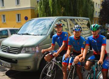 Trzech chłopców w sportowych strojach siedzi na rowerach przed zaparkowanym busem.