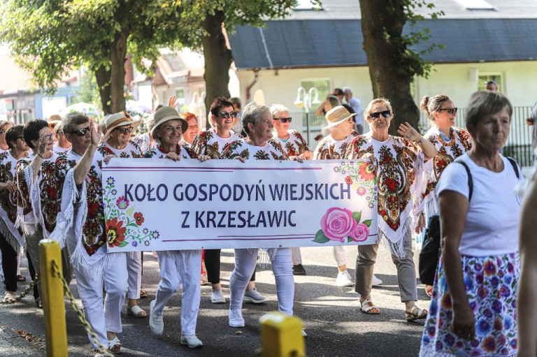 Grupa kobiet idzie w korowodzie. Panie mają wzorzyste czerwono białe chusty. Kobiety niosą baner.