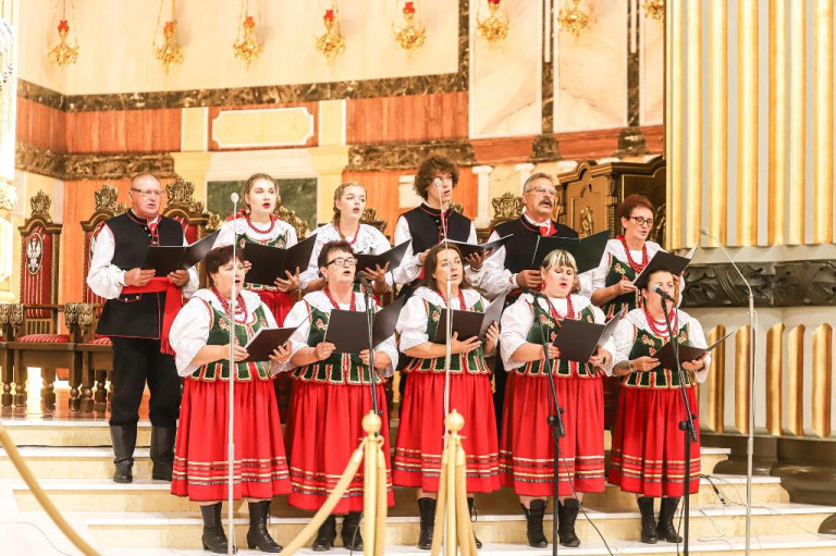 Grupa osób w ludowych strojach stoi przy ołtarzu i śpiewa.