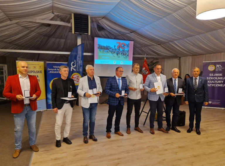Sala. Ośmiu mężczyzn stoi obok siebie w rzędzie. Siedmiu z nich trzyma medale i dyplomy.
