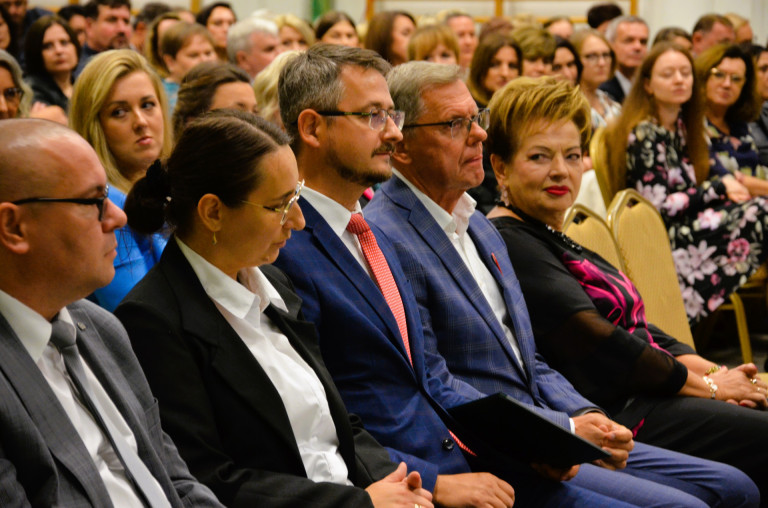 Zaproszeni goście siedzą na żółtych krzesłach.