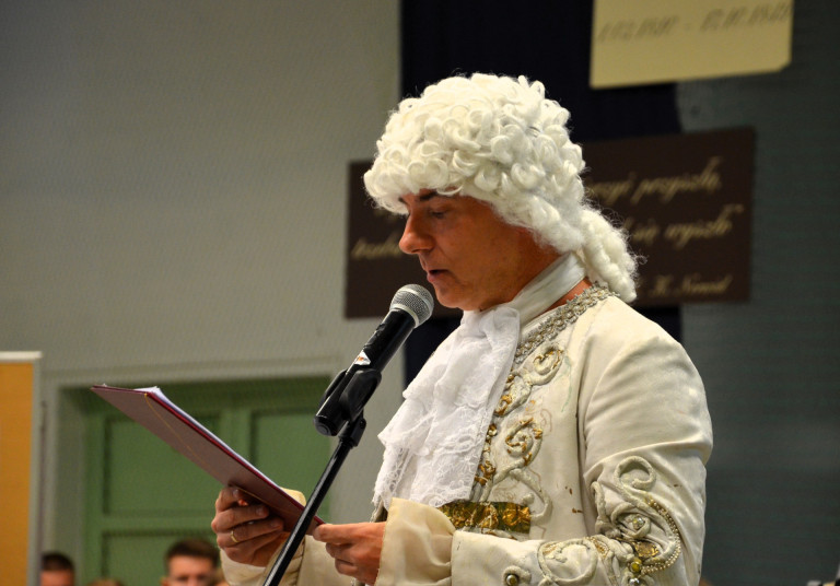 Mężczyzna w białej peruce i białym stroju z epoki oświecenia stoi i mówi do mikrofonu.