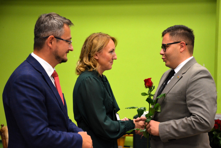 Dwóch mężczyzn w garniturach i kobieta w zielonym stroju. Kobieta odbiera gratulacje.