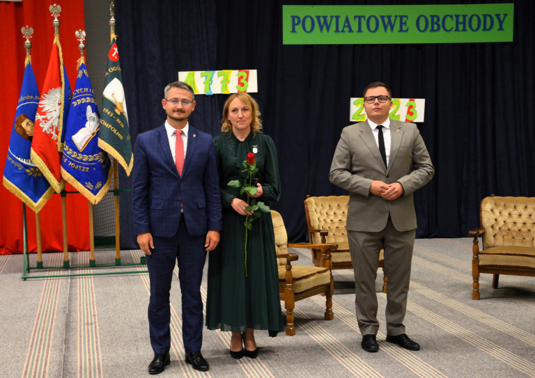 Dwóch mężczyzn w garniturach i kobieta w zielonym stroju. Kobieta ma przypięte do sukni odznaczenie.