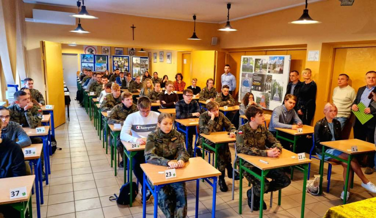 Uczestnicy konkursu siedzą w ławkach. Ławki ustawione są w czterech rzędach.