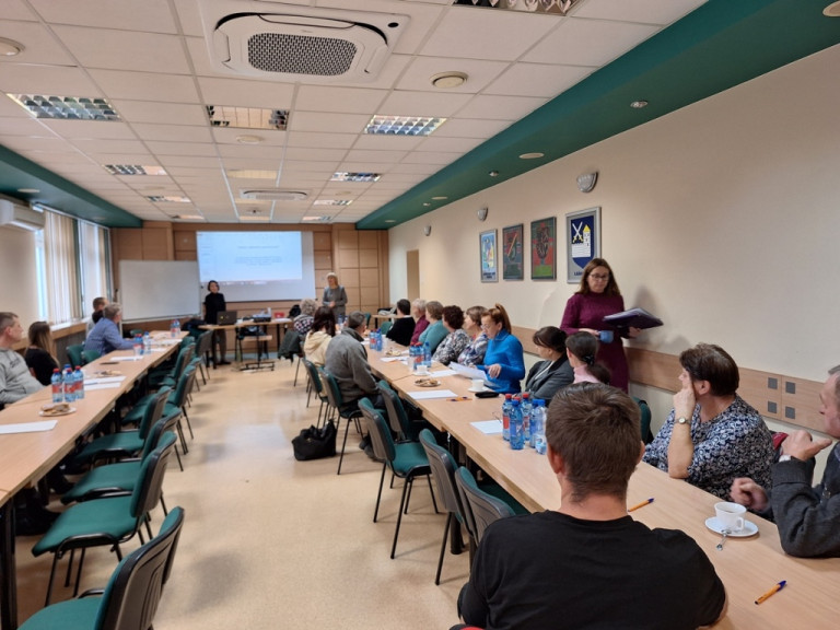 W sali siedzą uczestnicy spotkania. Dwie kobiety stoją przed ekranem projekcyjnym.