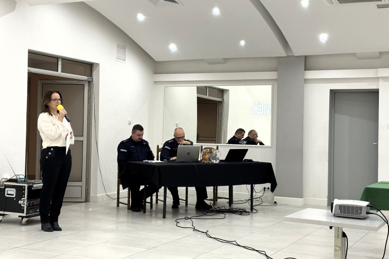 Kobieta w białej koszuli i czarnych spodniach mówi do mikrofonu. Dwóch polcjantów siedzi przy stole.