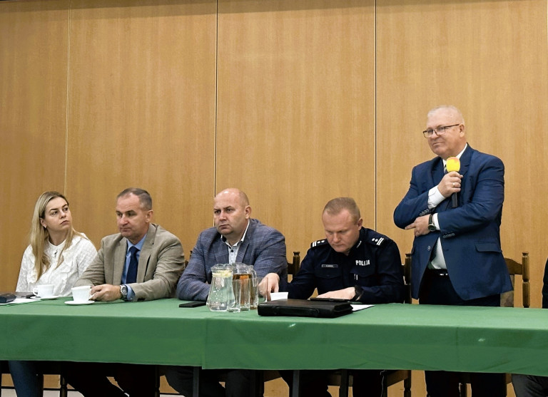 Kobieta i trzech mężczyzn siedzą przy stole. Jeden mężczyzna stoi i mówi do mikrofonu.