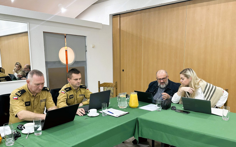 Dwóch strażaków oraz mężczyzna i kobieta siedzą przed laptopami.