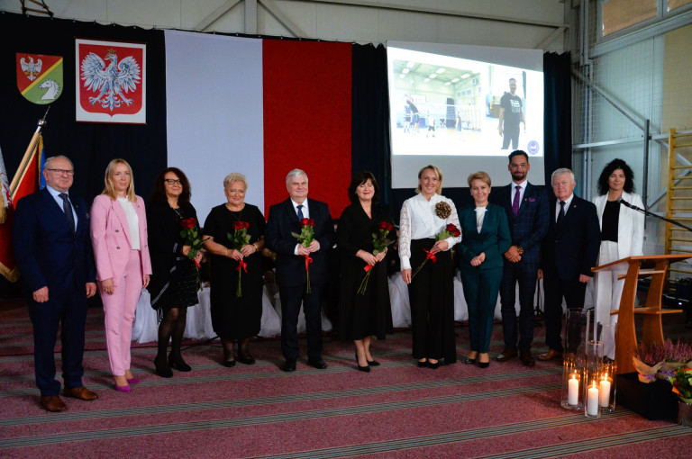 Grupa ludzi stoi na tle flagi i godła. Część z nich trzyma róże.