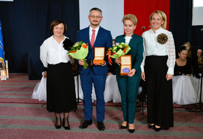 Czworo ludzi stoi na tle flagi. Dwoje z nich trzyma bukiety kwiatów.