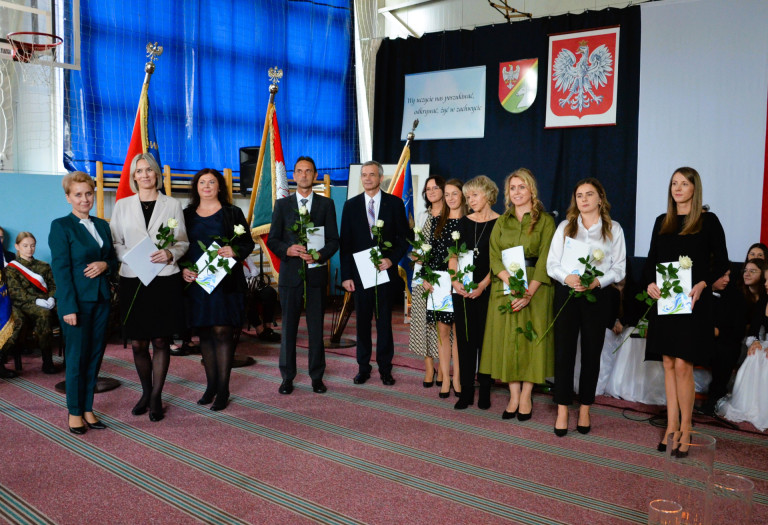 Grupa ludzi stoi na tle godła trzymając róże i dokumenty.