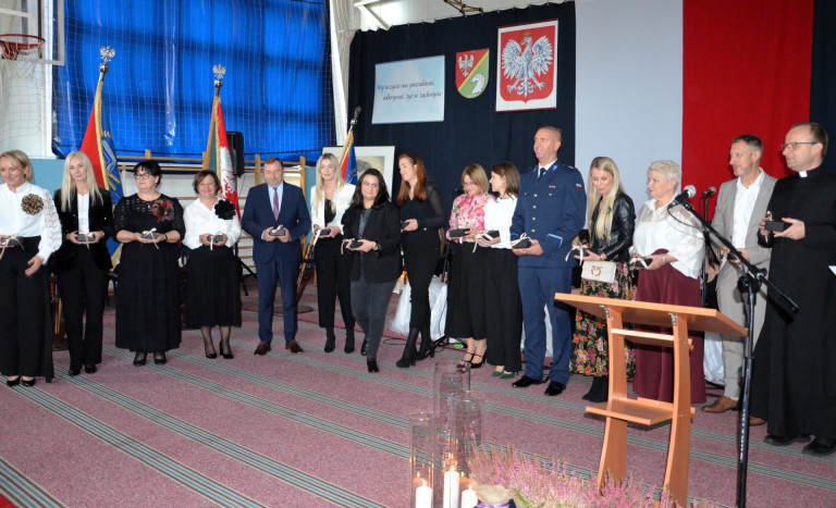Grupa ludzi stoi na tle godła trzymając upominki.