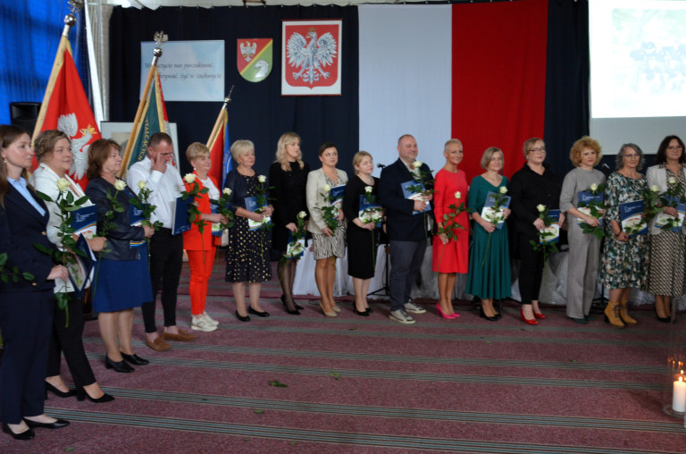 Grupa ludzi stoi na tle godła trzymając róże i dokumenty.