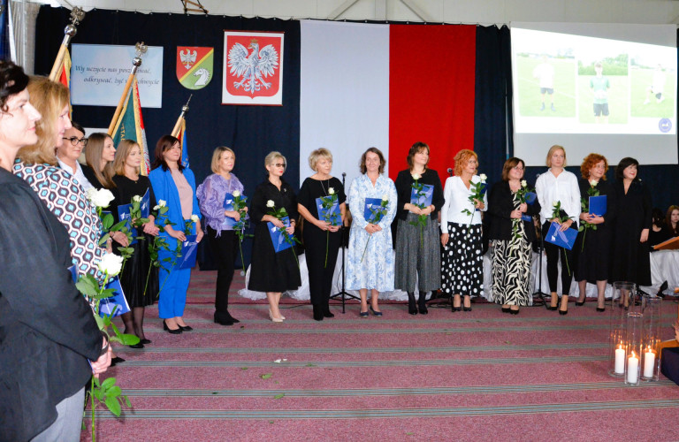 Grupa kobiet stoi na tle godła trzymając róże i dokumenty.
