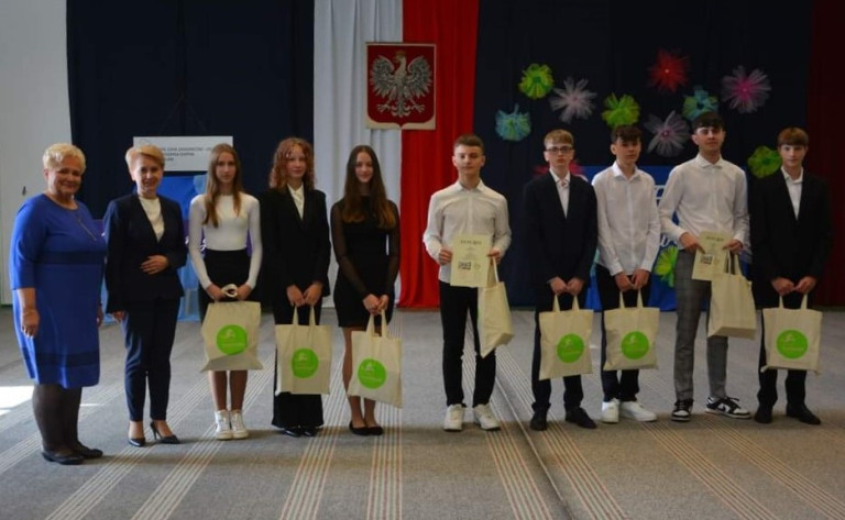 Grupa ludzi stoi na scenie. Część z nich trzyma upominki.