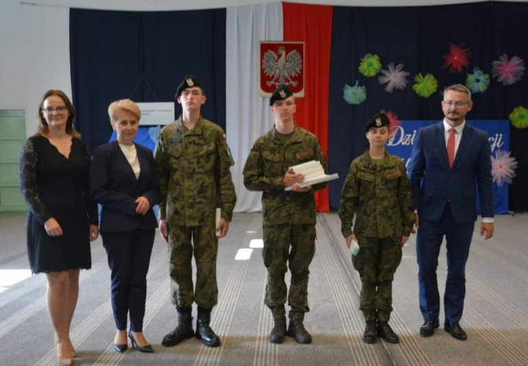 Grupa ludzi stoi w sali gimnastycznej na tle godła. Część z nich ubrana jest w mundury.
