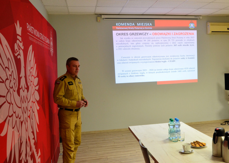 Strażak w żółtym mundurze przemawia w rogu sali.