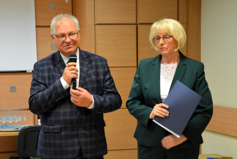 Kobieta w zielonej marynarce trzyma dokument. Obok niej mężczyzna przemawia do mikrofonu.