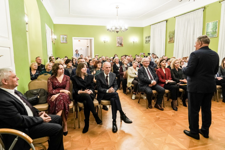 Widownia siedzi w zielonej sali. Przed nimi przemawia mężczyzna w garniturze.
