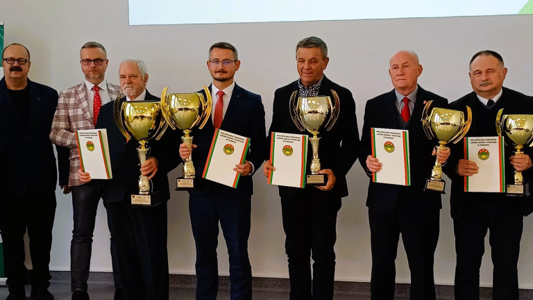 Grupa mężczyzn stoi na scenie. Część z nich trzyma złote puchary.