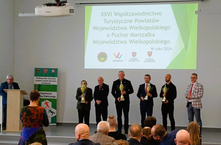 Grupa mężczyzn stoi na scenie. Część z nich trzyma złote puchary.