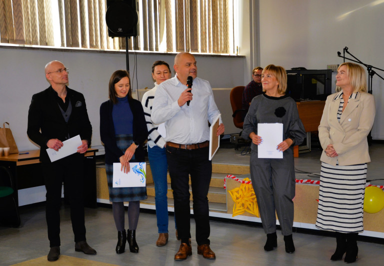 Grupa ludzi stoi przed sceną. Częśc z nich trzyma teczki.