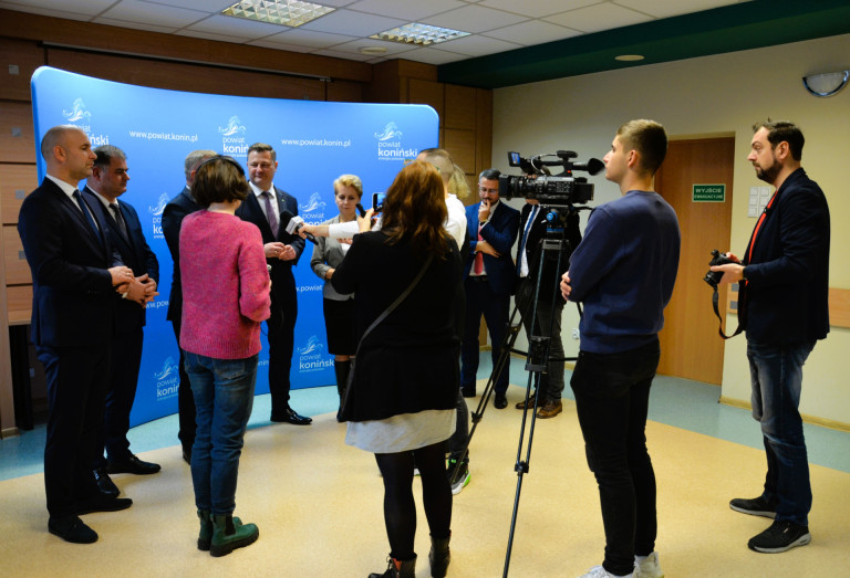 Grupa ludzi stoi otoczona mikrofonami dziennikarzy. Przed nimi stoi kamera.