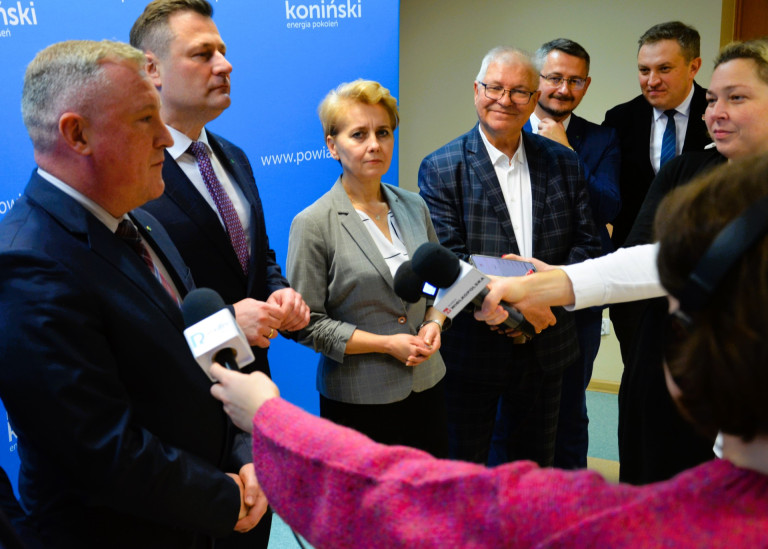 Grupa ludzi stoi otoczona mikrofonami dziennikarzy.