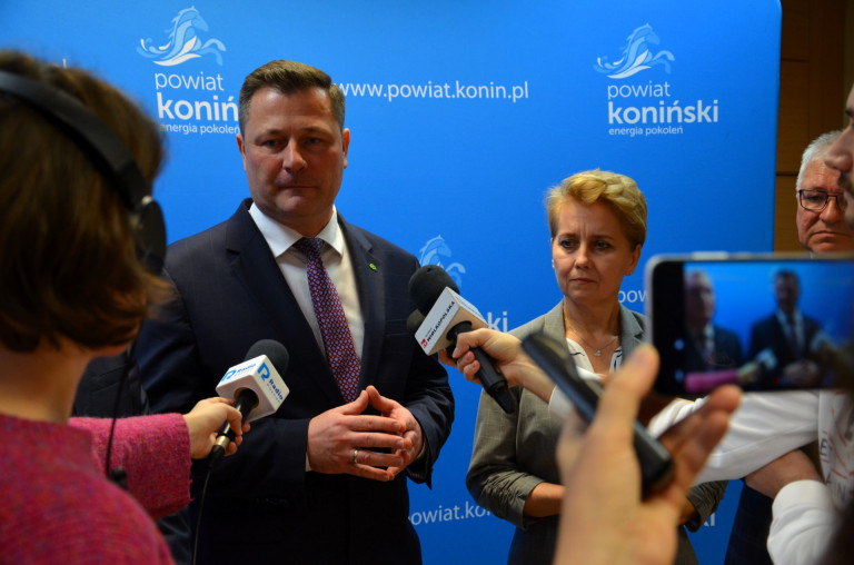 Kobieta i mężczyzna stoją na tle plakatu. Mężczyzna ma na siebie skierowane mikrofony.