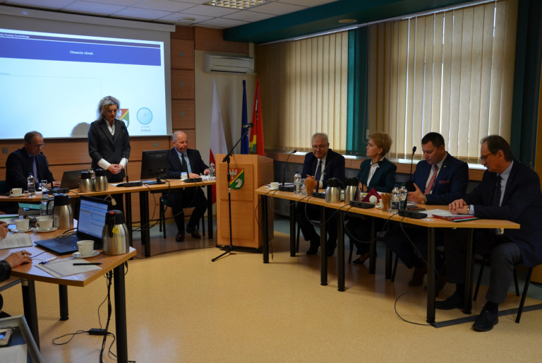 Grupa ludzi siedzi przy stołach w sali konferencyjnej. Jedna z kobiet przemawia stojąc za stołem.