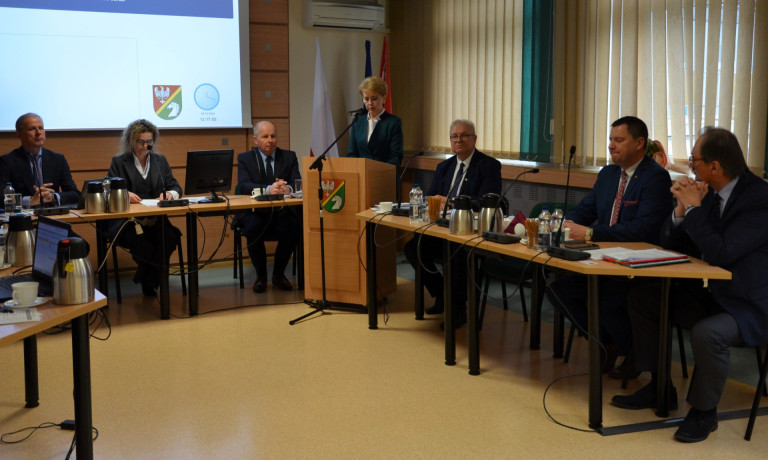 Grupa ludzi siedzi przy stołach w sali konferencyjnej. Kobieta w garniturze przemawia do mikrofonu.