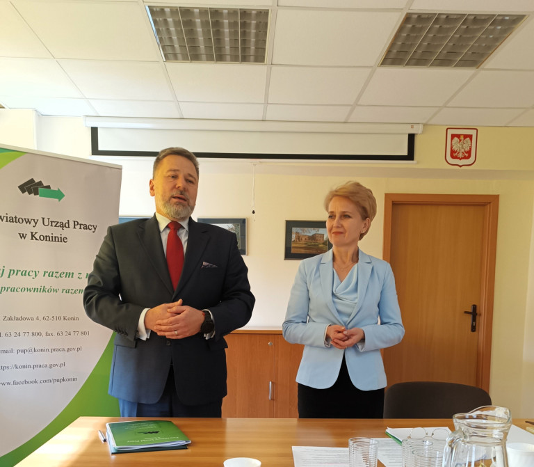 Kobieta w błękitnej marynarce  i mężczyzna w garniturze stoją za biurkiem.