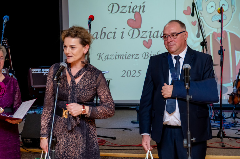 Mężczyzna i kobieta stoją przed sceną. Kobieta przemawia do mikrofonu.