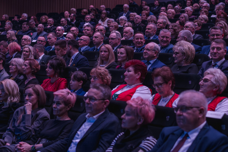 Liczna widownia siedzi w sali oświetlonej na różowo.