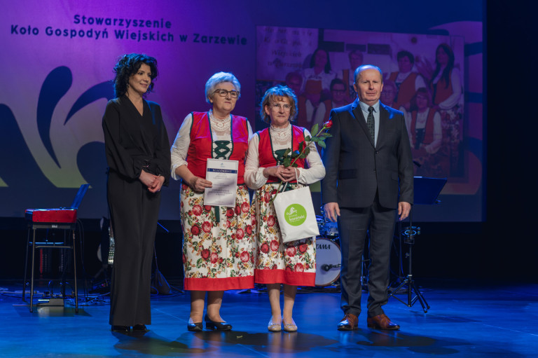 Trzy kobiety oraz mężczyzna stoją na scenie. Kobiety trzymają różę oraz nagrody.