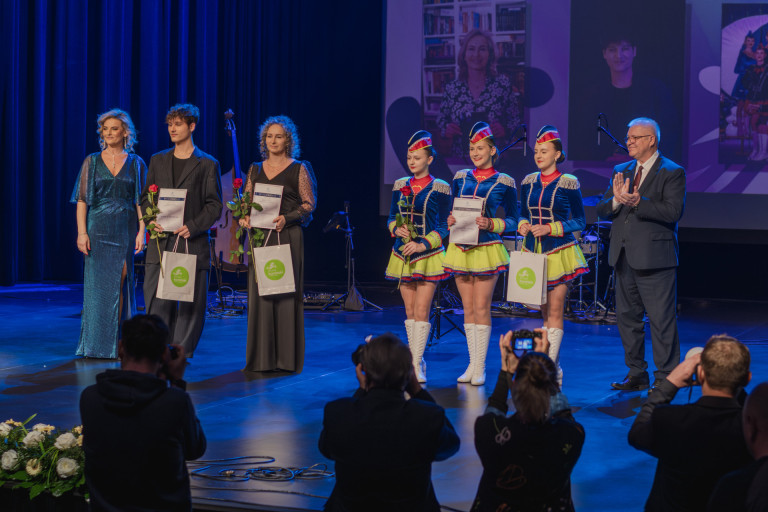 Trzy mażoretki oraz grupa ludzi stoi na scenie.