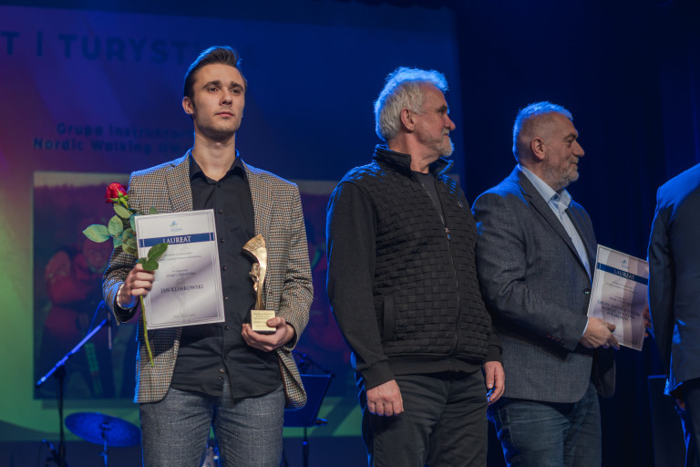 Trzech mężczyzn stoi na scenie. Jeden z nich trzyma statuetkę, różę i dyplom.