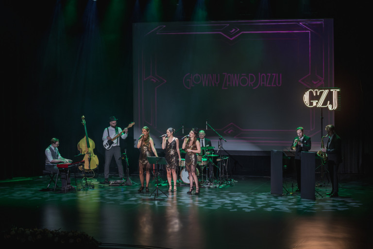Trzy kobiety występują na scenie. Kilku mężczyzn gra na instrumentach.
