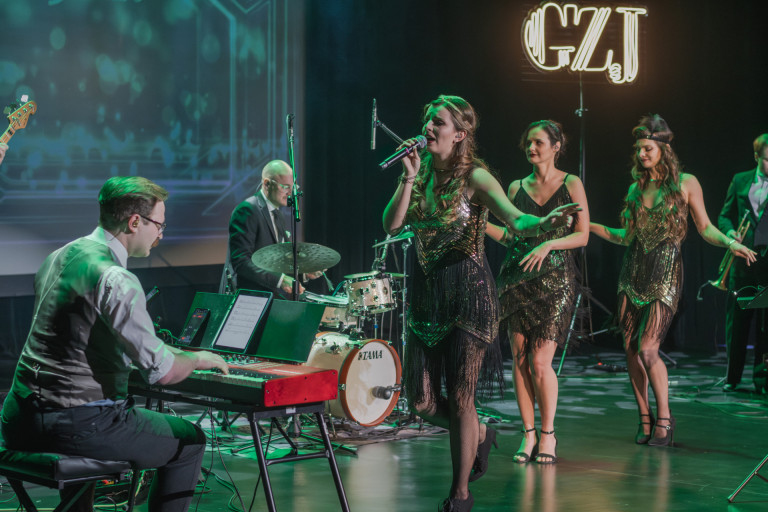 Trzy kobiety występują na scenie. Kilku mężczyzn gra na instrumentach.