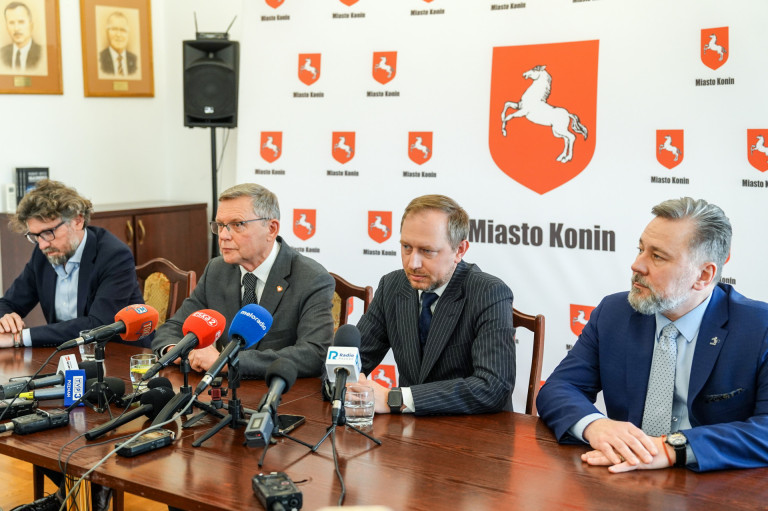 Czterech mężczyzn siedzi przy mikrofonach na tle baneru.