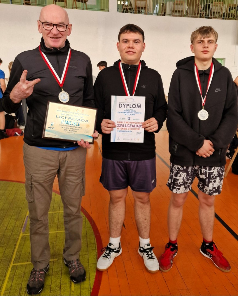 Trzech mężczyzn stoi w sali gimnastycznej. Każdy z nich ma medal na szyi.