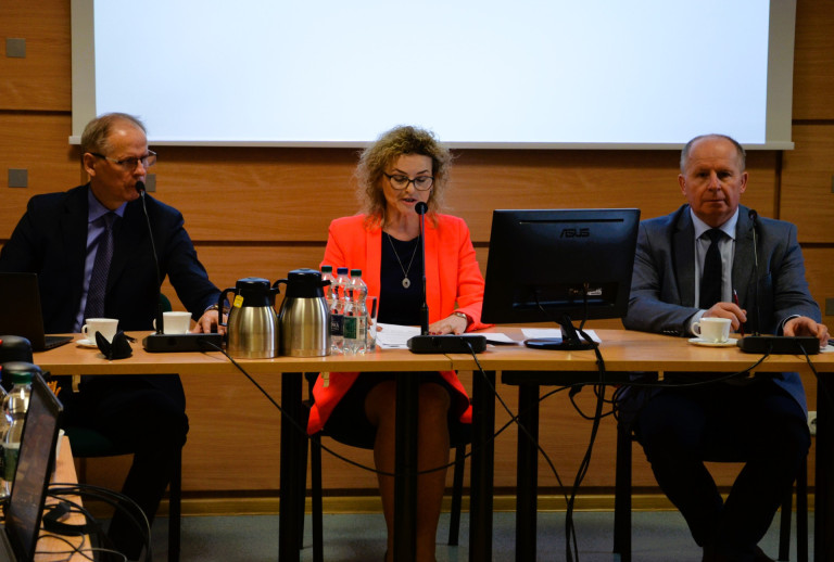 Dwóch mężczyzn oraz kobieta siedzą przy stole w sali konferencyjnej.