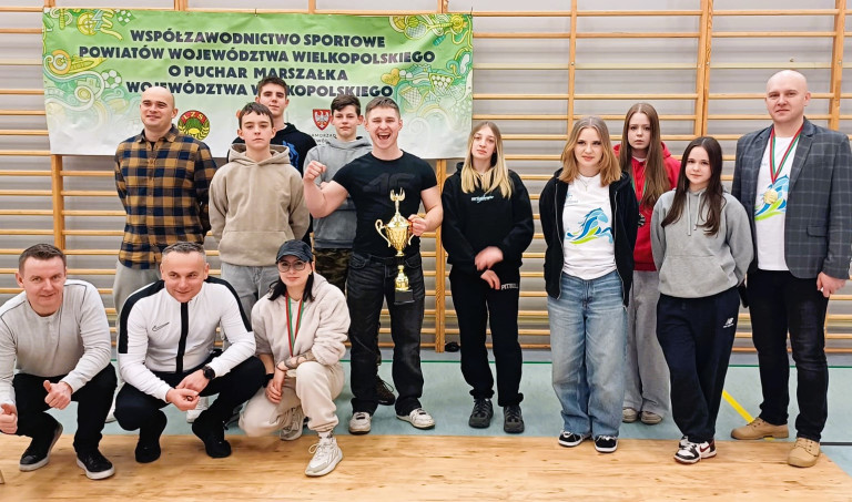 Kilkunastu ludzi stoi w sali gimnastycznej. Mężczyzna na środku trzyma złoty puchar.