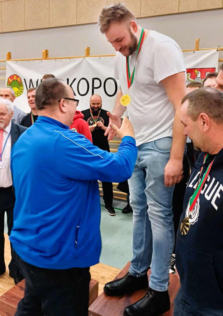 Mężczyzna stojący na podium podaje rękę mężczyźnie w niebieskiej kurtce.