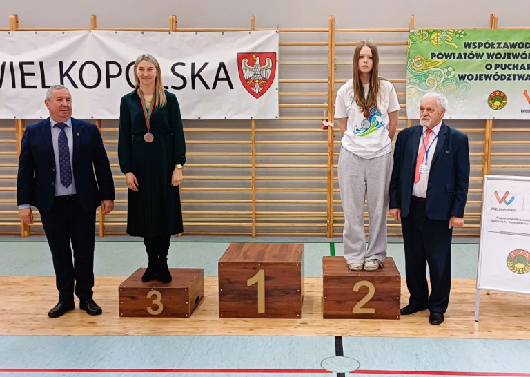Dwie kobiety stoją na podium. Obok podium stoi dwóch mężczyzn.