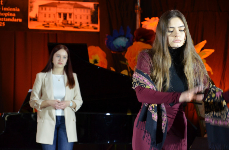 Kobieta stoi na scenie. Za nią stoi kobieta w kremowej marynarce.
