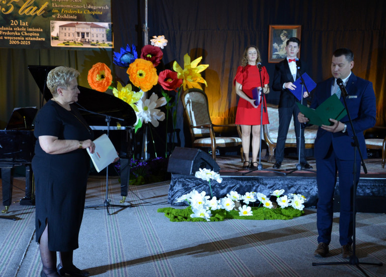 Mężczyzna w garniturze przemawia na scenie. W tle ozdoby jubileuszowe.
