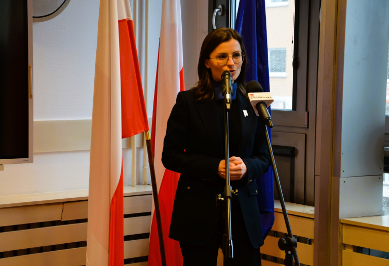 Kobieta w czarnym garniturze przemawia na tle flag.