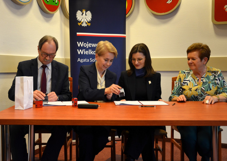 Trzy kobiety i mężczyzna siedzą przy stole. Jedna z kobiet oraz mężczyzna stawiają pieczątki.